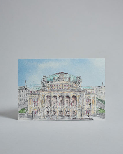 Die Vorderseite der Postkarte im Querformat, gedruckt auf hochwertigem Papier. Diese zeigt die Wiener Staatsoper vor blauem Himmel gemalt von einer Wiener Künstlerin. Links neben dem Gebäude ist der Schriftzug "Vienna" in dezenter, geschwungener Schrift zu lesen.