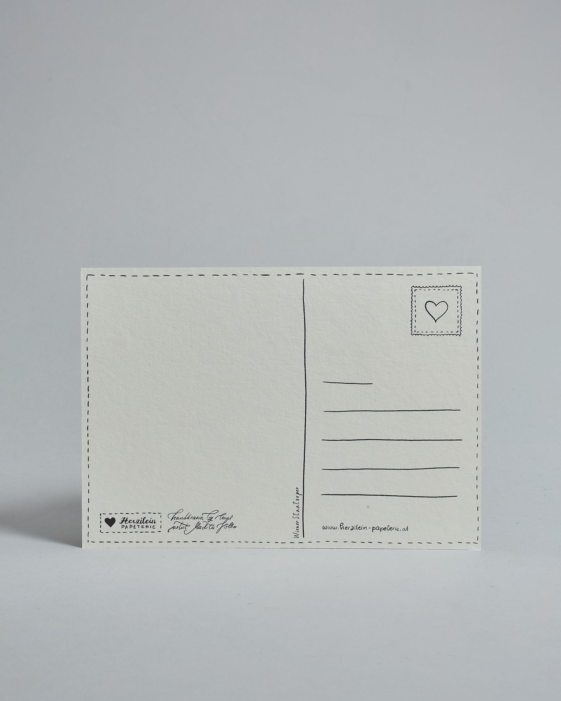 Die Rückseite der Postkarte im Querformat vor hellgrauem Hintergrund. 
Ein handgezeichnetes, Postkarten-typisches Layout mit Adressfeld auf der rechten Seite und Textfeld auf der linken Seite ist zu sehen, dazwischen eine Trennlinie. Auf der Trennlinie steht der auf der Vorderseite dargestellte Ort.  Rechts oben ist eine stilisiere Briefmarke mit Herz-Motiv zu sehen. Links unten steht das Logo Herzilein-Papeterie und daneben der Text: "handdrawn by local artist Madita Völker".