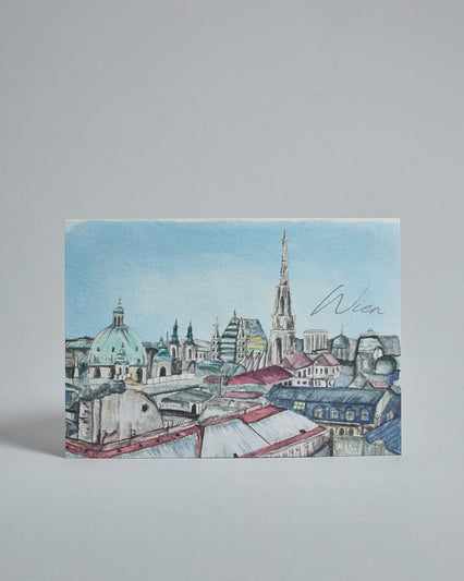 Die Vorderseite der Postkarte im Querformat, gedruckt auf hochwertigem Papier. Diese zeigt die Dächer über der Wiener Innenstadt gemalt von einer Wiener Künstlerin. Bekannte erkennbare Gebäude sind der Stephansdom und die Kuppel der Peterskirche. Rechts neben dem Stephansdom ist "Wien" in dezenter geschwungener Schrift zu lesen.