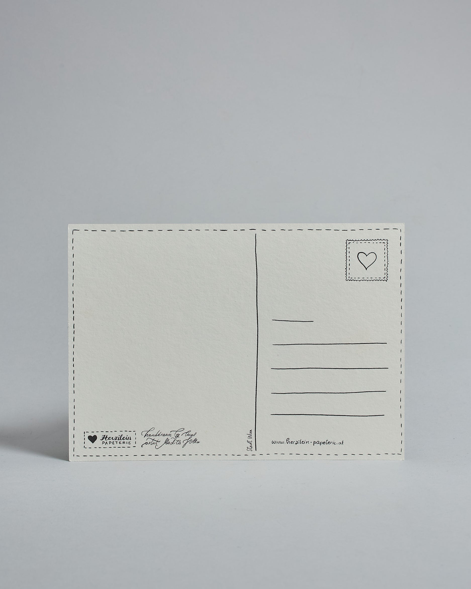 Die Rückseite der Postkarte im Querformat vor hellgrauem Hintergrund. 
Ein handgezeichnetes, Postkarten-typisches Layout mit Adressfeld auf der rechten Seite und Textfeld auf der linken Seite ist zu sehen, dazwischen eine Trennlinie. Auf der Trennlinie steht der auf der Vorderseite dargestellte Ort.  Rechts oben ist eine stilisiere Briefmarke mit Herz-Motiv zu sehen. Links unten steht das Logo Herzilein-Papeterie und daneben der Text: "handdrawn by local artist Madita Völker".