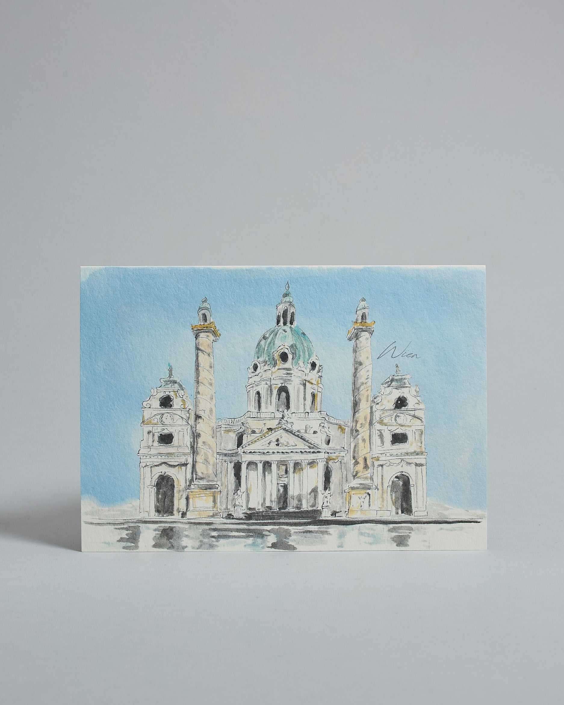 Die Vorderseite der Postkarte im Querformat, gedruckt auf hochwertigem Papier. Diese zeigt die Wiener Karlskirche vor blauem Himmel gemalt von einer Wiener Künstlerin. Im unteren Teil der Karte ist ein Brunnen zu sehen in dem sich die Kirche spiegelt.
Rechts neben der Kirche ist "Wien" in dezenter, geschwungener Schrift zu lesen.