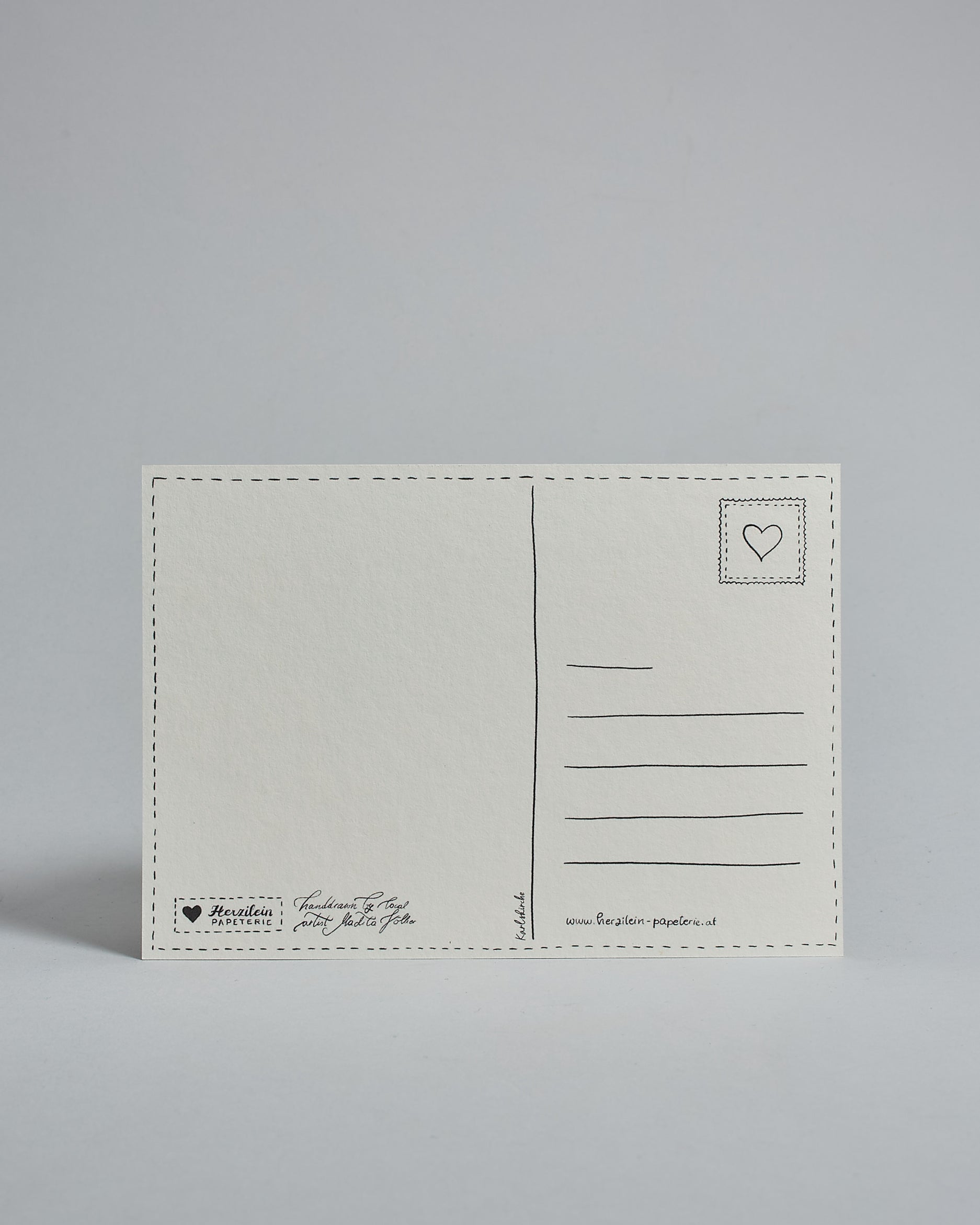 Die Rückseite der Postkarte im Querformat vor hellgrauem Hintergrund. 
Ein handgezeichnetes, Postkarten-typisches Layout mit Adressfeld auf der rechten Seite und Textfeld auf der linken Seite ist zu sehen, dazwischen eine Trennlinie. Auf der Trennlinie steht der auf der Vorderseite dargestellte Ort.  Rechts oben ist eine stilisiere Briefmarke mit Herz-Motiv zu sehen. Links unten steht das Logo Herzilein-Papeterie und daneben der Text: "handdrawn by local artist Madita Völker".