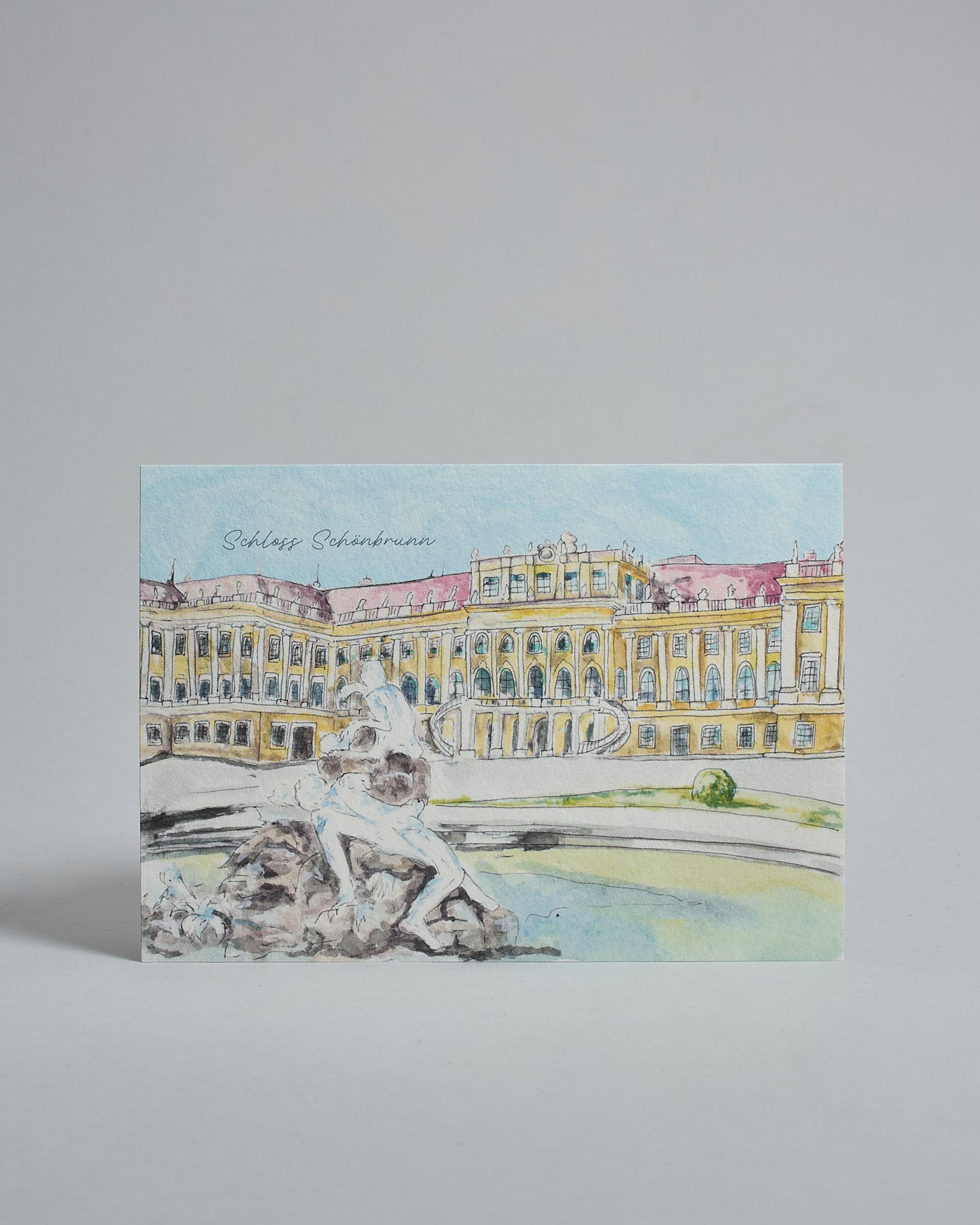 Die Vorderseite der Postkarte im Querformat, gedruckt auf hochwertigem Papier. Diese zeigt das Wiener Schloss Schönbrunn von seiner Vorderseite, gemalt von einer Wiener Künstlerin. Im Vordergrund ist außerdem ein Brunnen zu sehen. Links über dem Schloss ist "Schloss Schönbrunn" in dezenter, geschwungener Schrift zu lesen.