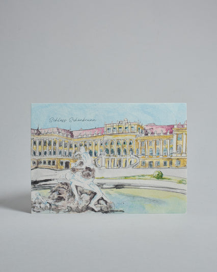 Die Vorderseite der Postkarte im Querformat, gedruckt auf hochwertigem Papier. Diese zeigt das Wiener Schloss Schönbrunn von seiner Vorderseite, gemalt von einer Wiener Künstlerin. Im Vordergrund ist außerdem ein Brunnen zu sehen. Links über dem Schloss ist "Schloss Schönbrunn" in dezenter, geschwungener Schrift zu lesen.