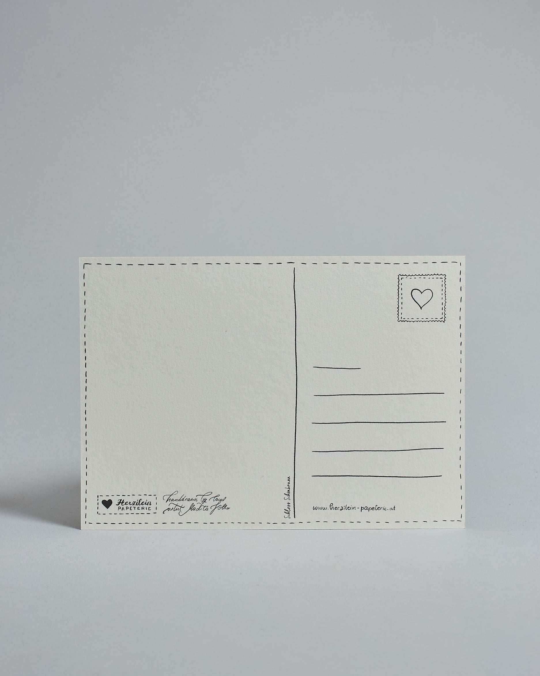 Die Rückseite der Postkarte im Querformat vor hellgrauem Hintergrund. 
Ein handgezeichnetes, Postkarten-typisches Layout mit Adressfeld auf der rechten Seite und Textfeld auf der linken Seite ist zu sehen, dazwischen eine Trennlinie. Auf der Trennlinie steht der auf der Vorderseite dargestellte Ort.  Rechts oben ist eine stilisiere Briefmarke mit Herz-Motiv zu sehen. Links unten steht das Logo Herzilein-Papeterie und daneben der Text: "handdrawn by local artist Madita Völker".