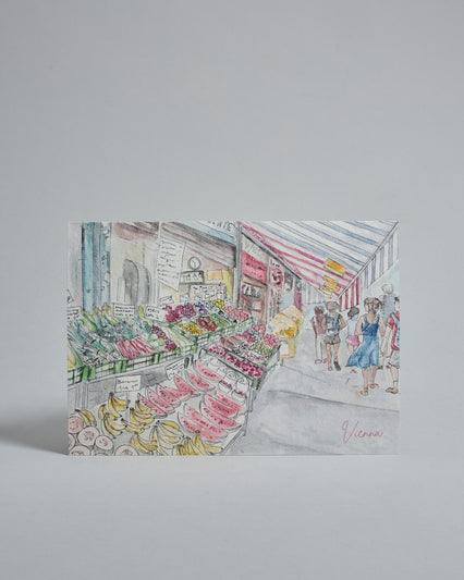 Die Vorderseite der Postkarte im Querformat, gedruckt auf hochwertigem Papier. Diese zeigt den Wiener Naschmarkt gemalt von einer Wiener Künstlerin. Zu sehen ist ein Obst mit gestreifter Markise und einigen Passanten im rechten Teil der Karte. Rechts unten ist "Vienna" in dezenter, geschwungener Schrift zu lesen.
