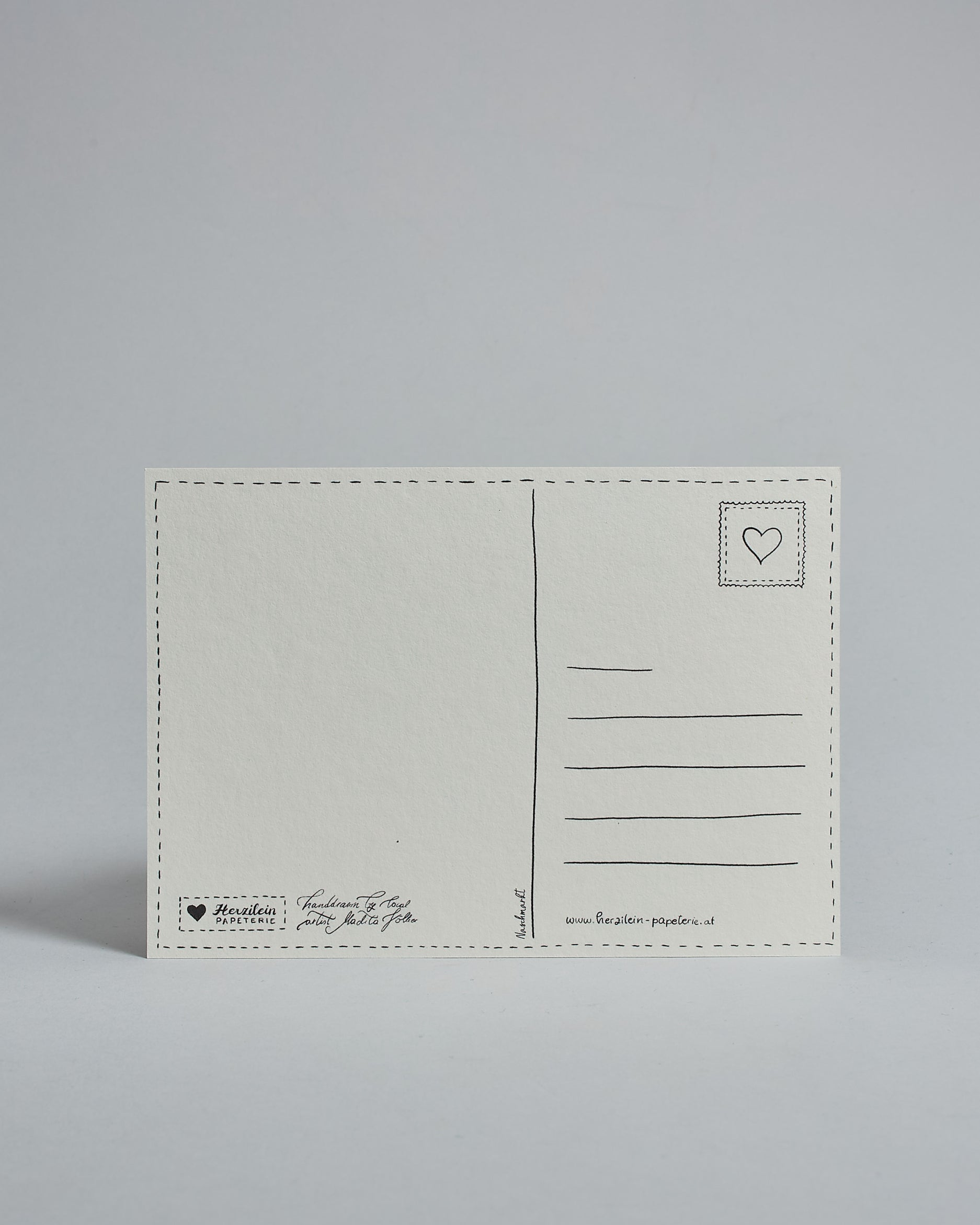 Die Rückseite der Postkarte im Querformat vor hellgrauem Hintergrund. 
Ein handgezeichnetes, Postkarten-typisches Layout mit Adressfeld auf der rechten Seite und Textfeld auf der linken Seite ist zu sehen, dazwischen eine Trennlinie. Auf der Trennlinie steht der auf der Vorderseite dargestellte Ort.  Rechts oben ist eine stilisiere Briefmarke mit Herz-Motiv zu sehen. Links unten steht das Logo Herzilein-Papeterie und daneben der Text: "handdrawn by local artist Madita Völker".