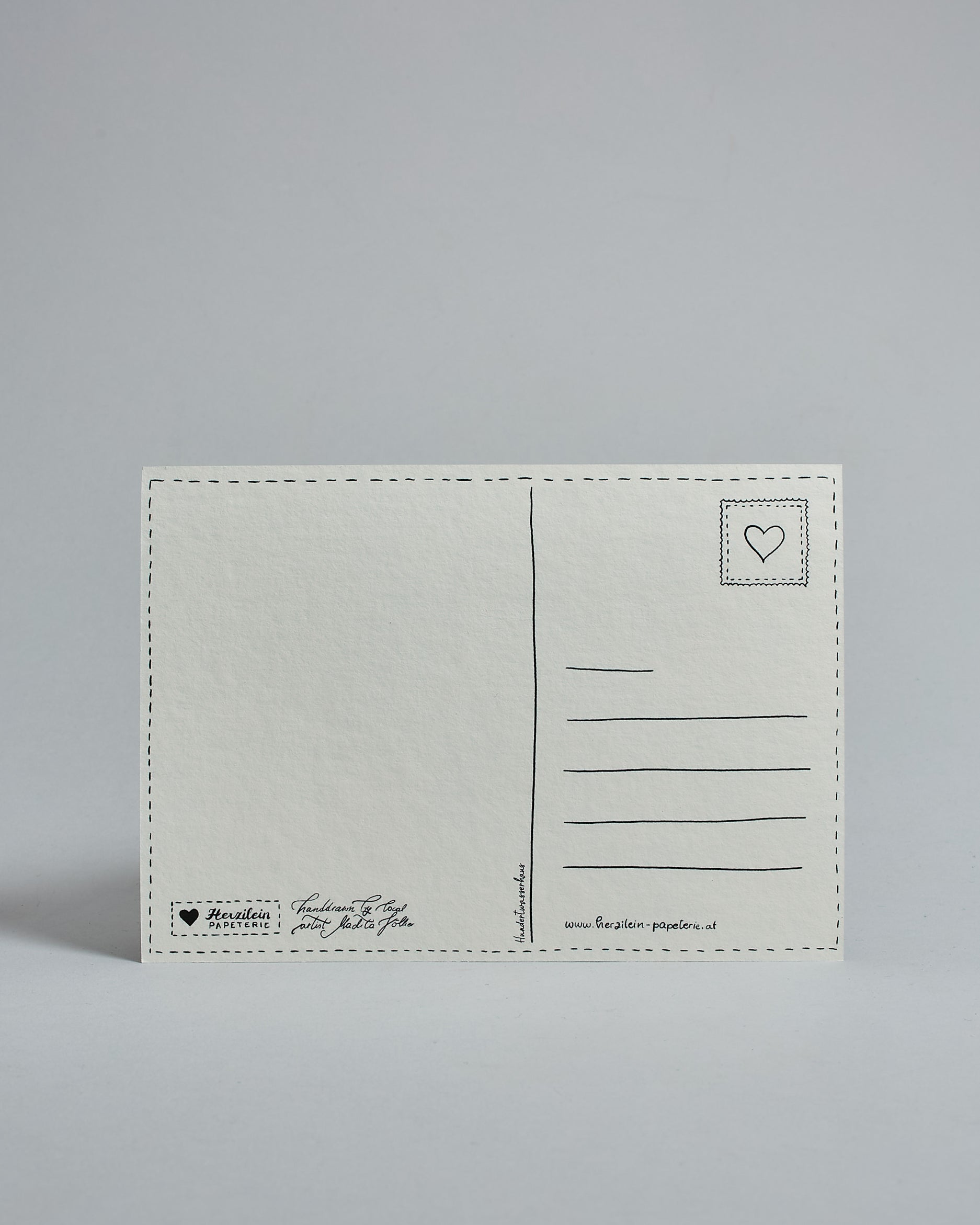 Die Rückseite der Postkarte im Querformat vor hellgrauem Hintergrund. 
Ein handgezeichnetes, Postkarten-typisches Layout mit Adressfeld auf der rechten Seite und Textfeld auf der linken Seite ist zu sehen, dazwischen eine Trennlinie. Auf der Trennlinie steht der auf der Vorderseite dargestellte Ort.  Rechts oben ist eine stilisiere Briefmarke mit Herz-Motiv zu sehen. Links unten steht das Logo Herzilein-Papeterie und daneben der Text: "handdrawn by local artist Madita Völker".