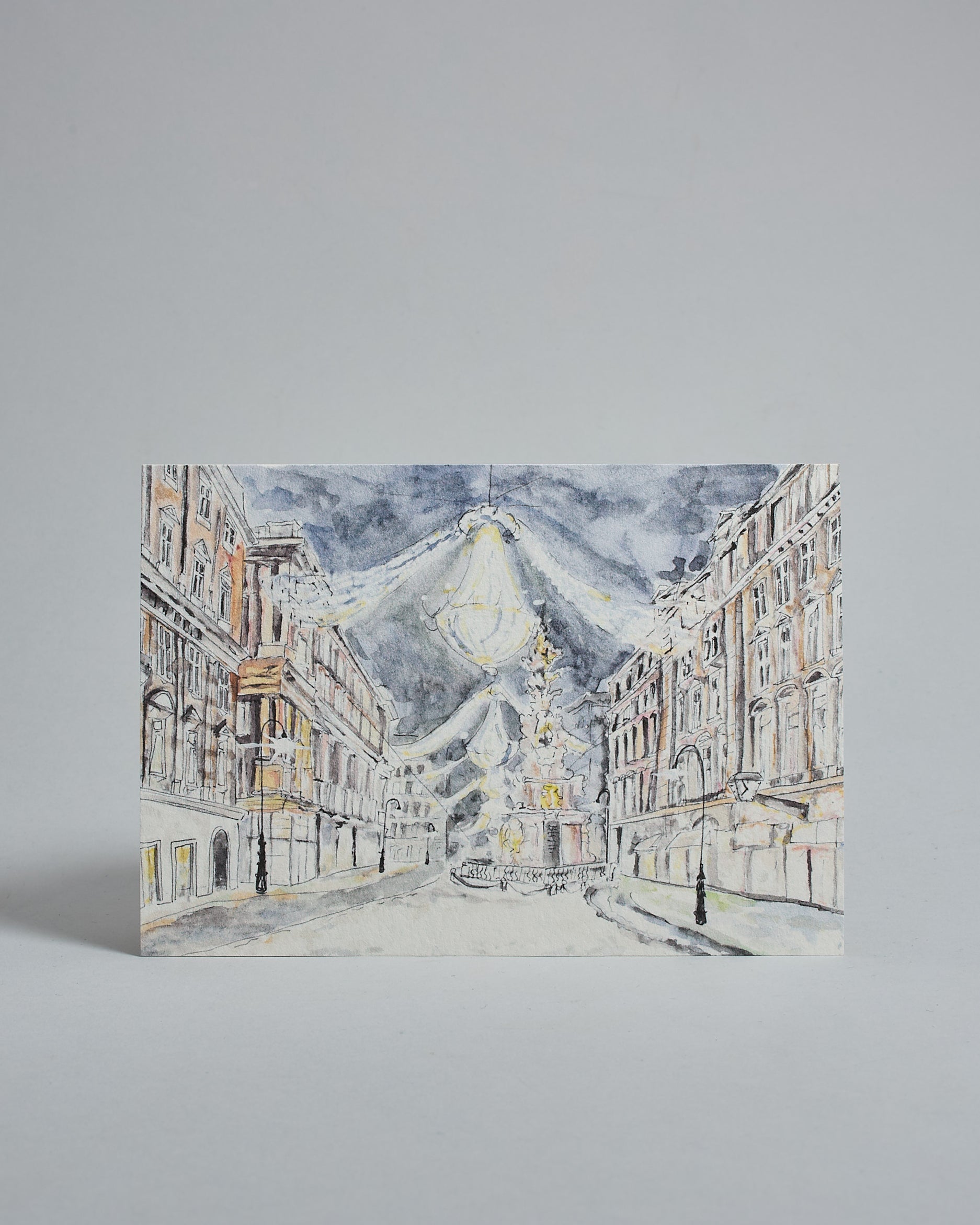 Die Vorderseite der Postkarte im Querformat, gedruckt auf hochwertigem Papier. Diese zeigt den Wiener Graben  mit Weihnachtsbeleuchtung gemalt von einer Wiener Künstlerin. Auch zu sehen ist die Pestsäule.
