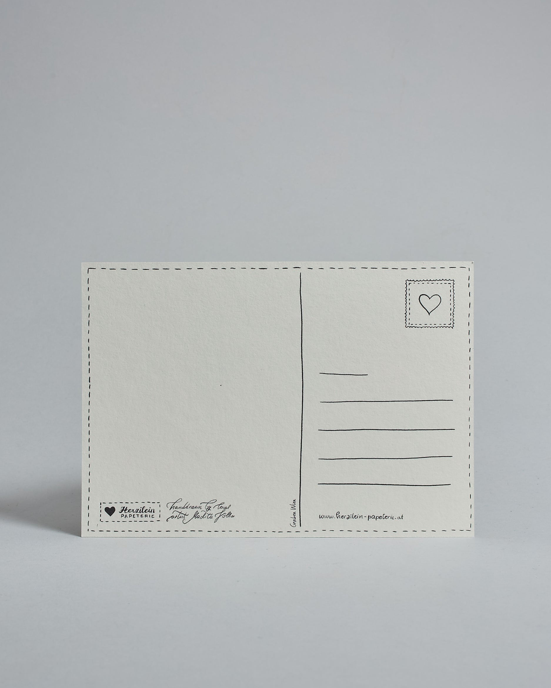 Die Rückseite der Postkarte im Querformat vor hellgrauem Hintergrund. 
Ein handgezeichnetes, Postkarten-typisches Layout mit Adressfeld auf der rechten Seite und Textfeld auf der linken Seite ist zu sehen, dazwischen eine Trennlinie. Auf der Trennlinie steht der auf der Vorderseite dargestellte Ort.  Rechts oben ist eine stilisiere Briefmarke mit Herz-Motiv zu sehen. Links unten steht das Logo Herzilein-Papeterie und daneben der Text: "handdrawn by local artist Madita Völker".