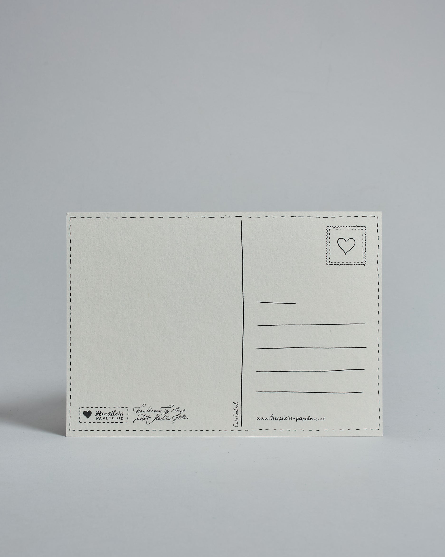 Die Rückseite der Postkarte im Querformat vor hellgrauem Hintergrund. 
Ein handgezeichnetes, Postkarten-typisches Layout mit Adressfeld auf der rechten Seite und Textfeld auf der linken Seite ist zu sehen, dazwischen eine Trennlinie. Auf der Trennlinie steht der auf der Vorderseite dargestellte Ort.  Rechts oben ist eine stilisiere Briefmarke mit Herz-Motiv zu sehen. Links unten steht das Logo Herzilein-Papeterie und daneben der Text: "handdrawn by local artist Madita Völker".