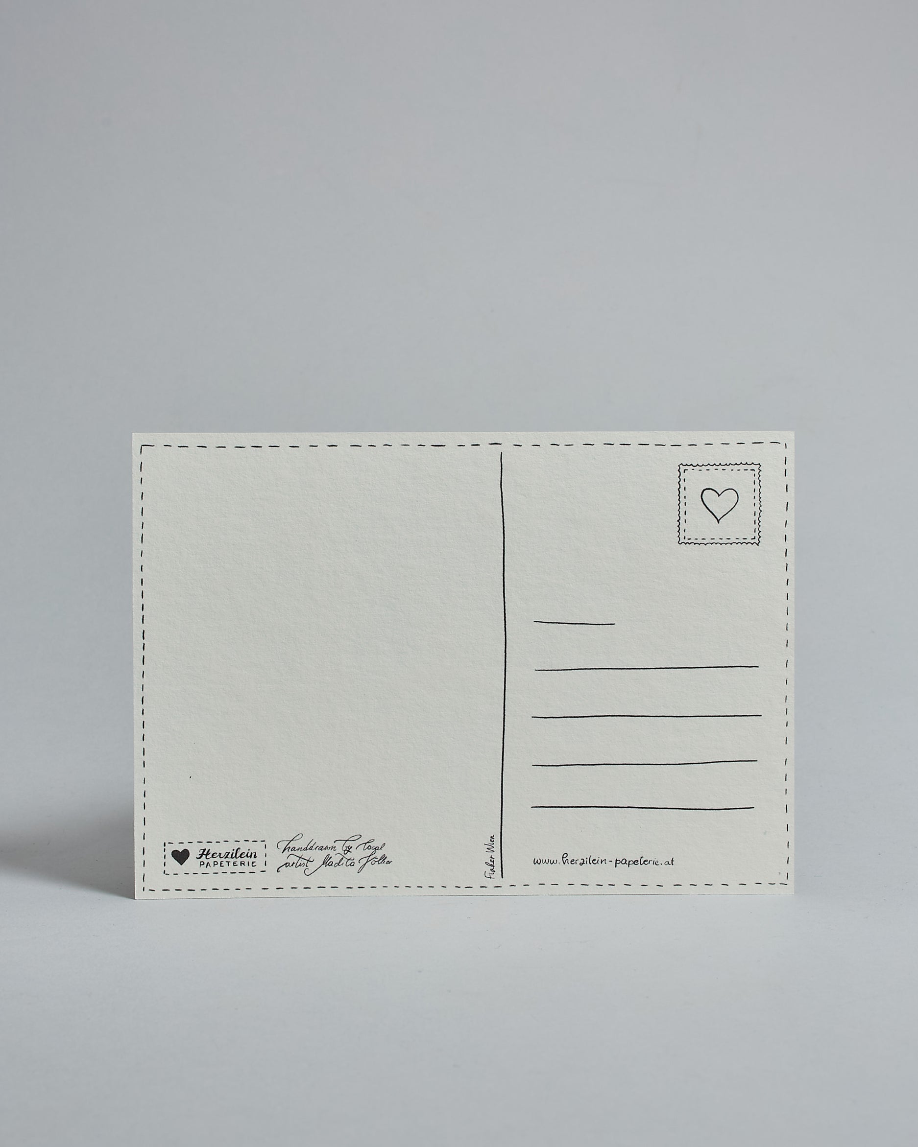 Die Rückseite der Postkarte im Querformat vor hellgrauem Hintergrund. 
Ein handgezeichnetes, Postkarten-typisches Layout mit Adressfeld auf der rechten Seite und Textfeld auf der linken Seite ist zu sehen, dazwischen eine Trennlinie. Auf der Trennlinie steht der auf der Vorderseite dargestellte Ort.  Rechts oben ist eine stilisiere Briefmarke mit Herz-Motiv zu sehen. Links unten steht das Logo Herzilein-Papeterie und daneben der Text: "handdrawn by local artist Madita Völker".