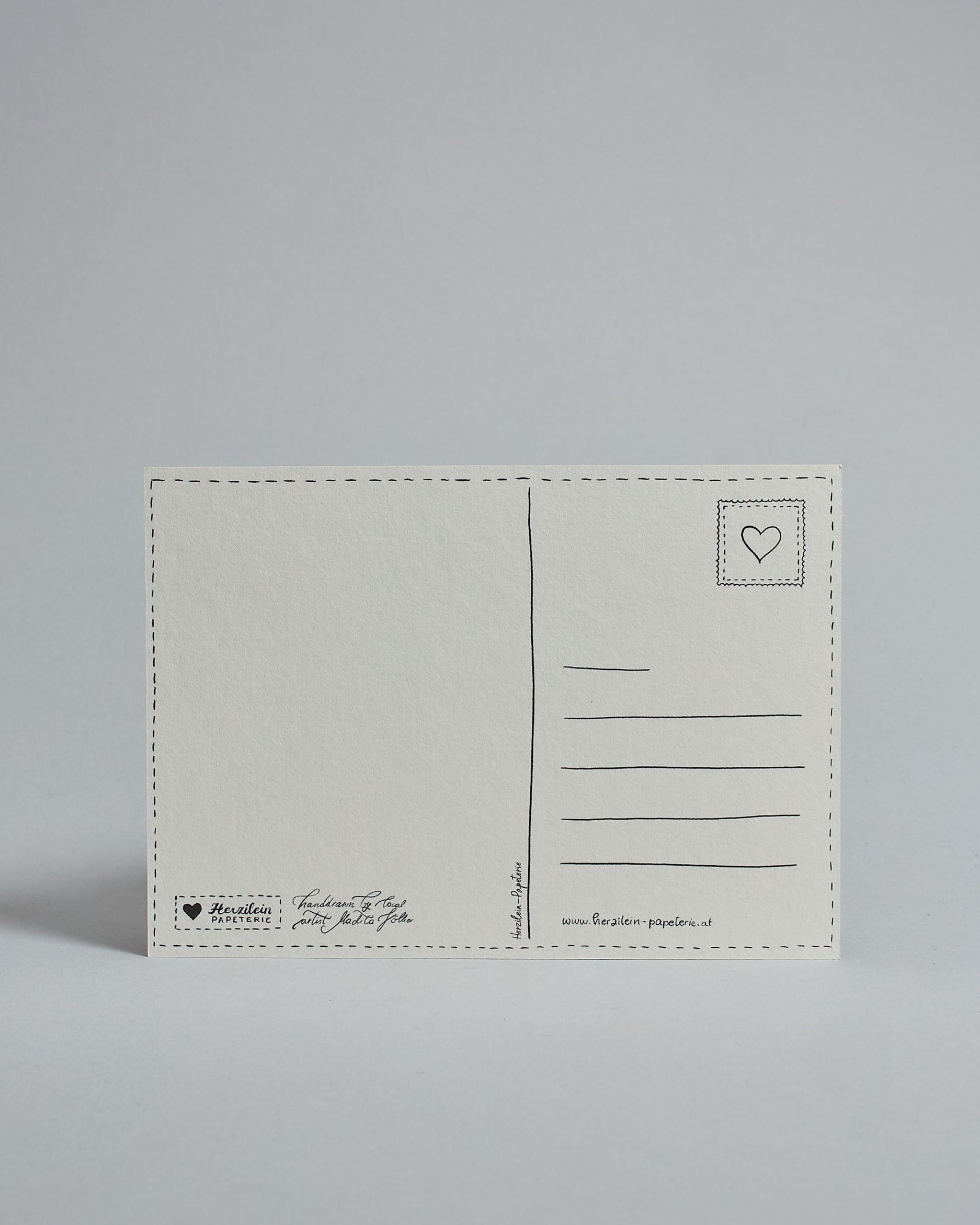 Die Rückseite der Postkarte im Querformat vor hellgrauem Hintergrund. 
Ein handgezeichnetes, Postkarten-typisches Layout mit Adressfeld auf der rechten Seite und Textfeld auf der linken Seite ist zu sehen, dazwischen eine Trennlinie. Auf der Trennlinie steht der auf der Vorderseite dargestellte Ort.  Rechts oben ist eine stilisiere Briefmarke mit Herz-Motiv zu sehen. Links unten steht das Logo Herzilein-Papeterie und daneben der Text: "handdrawn by local artist Madita Völker".