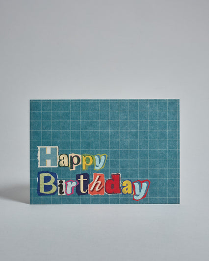 Die Vorderseite der Postkarte im Querformat vor hellgrauem Hintergrund. Im Vordergrund der Postkarte steht "Happy Birthday" in zwei Zeilen in Buchstaben die aussehen als wären sie aus Zeitrschriften ausgeschnitten. Der Hintergrund ist blau mit dezentem Karomuster.