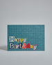 Die Vorderseite der Postkarte im Querformat vor hellgrauem Hintergrund. Im Vordergrund der Postkarte steht "Happy Birthday" in zwei Zeilen in Buchstaben die aussehen als wären sie aus Zeitrschriften ausgeschnitten. Der Hintergrund ist blau mit dezentem Karomuster.
