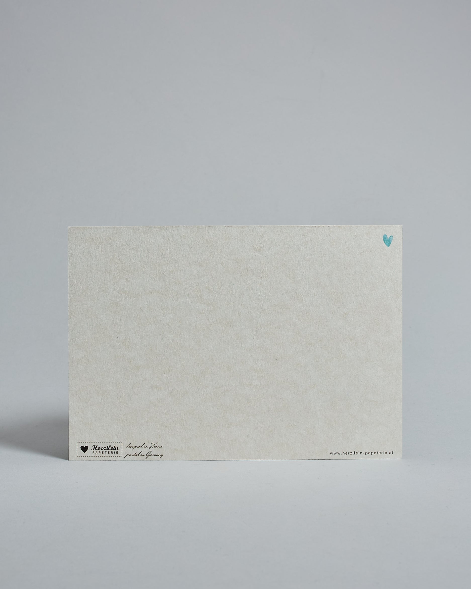 Die Rückseite der Postkarte im Querformat vor hellgrauem Hintergrund. Rechts oben ist, in kleiner Ausführung, ein hellblaues Herz zu sehen. 
Unten sind klein das Logo Herzilein-Papeterie, der Text: "designed in VIenna, printed in Germany" und "www.herzilein-papeterie.at" zu sehen. 
Der Großteil ist unbedruckt die Struktur der hochwertigen Holzschliffpappe ist gut zu sehen. Hier ist viel Platz zum Schreiben.