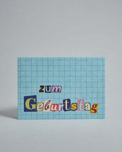 Die Vorderseite der Postkarte im Querformat vor hellgrauem Hintergrund. Im Vordergrund der Postkarte steht "zum Geburtstag" in zwei Zeilen in Buchstaben die aussehen als wären sie aus Zeitrschriften ausgeschnitten. Der Hintergrund ist hellblau mit dezentem Karomuster.