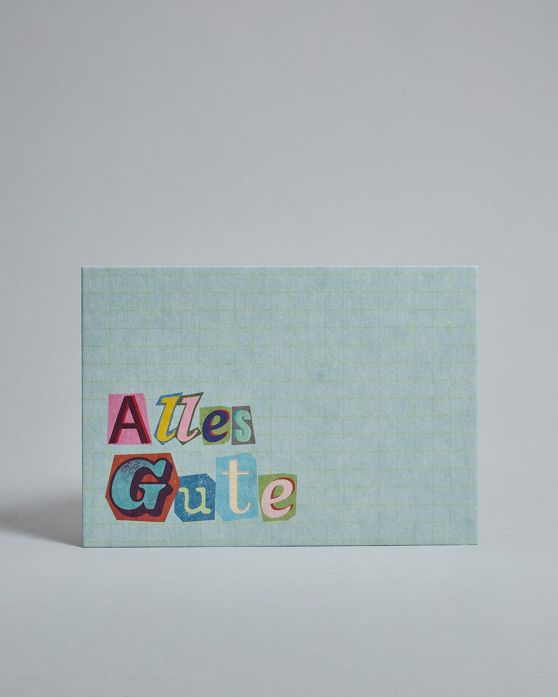 Die Vorderseite der Postkarte im Querformat vor hellgrauem Hintergrund. Im Vordergrund der Postkarte steht "Alles Gute" in Buchstaben die aussehen als wären sie aus Zeitrschriften ausgeschnitten. Der Hintergrund ist hellblau mit dezentem Karomuster.