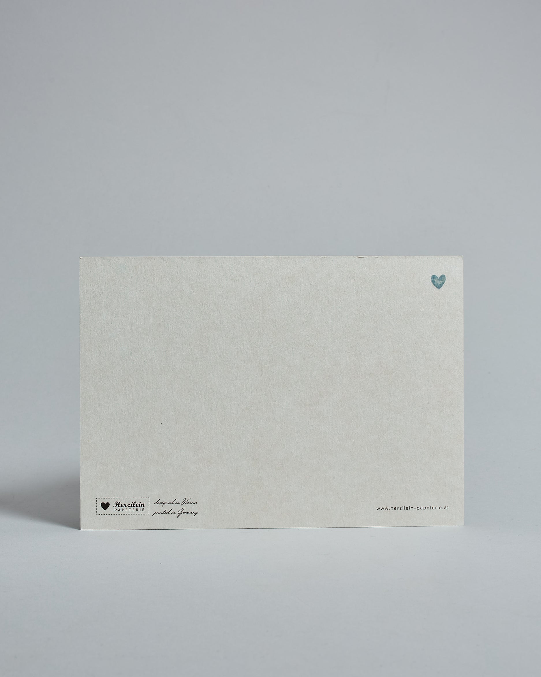 Die Rückseite der Postkarte im Querformat vor hellgrauem Hintergrund. Rechts oben ist, in kleiner Ausführung, ein blaues Herz zu sehen. 
Unten sind klein das Logo Herzilein-Papeterie, der Text: "designed in VIenna, printed in Germany" und "www.herzilein-papeterie.at" zu sehen. 
Der Großteil ist unbedruckt die Struktur der hochwertigen Holzschliffpappe ist gut zu sehen. Hier ist viel Platz zum Schreiben.