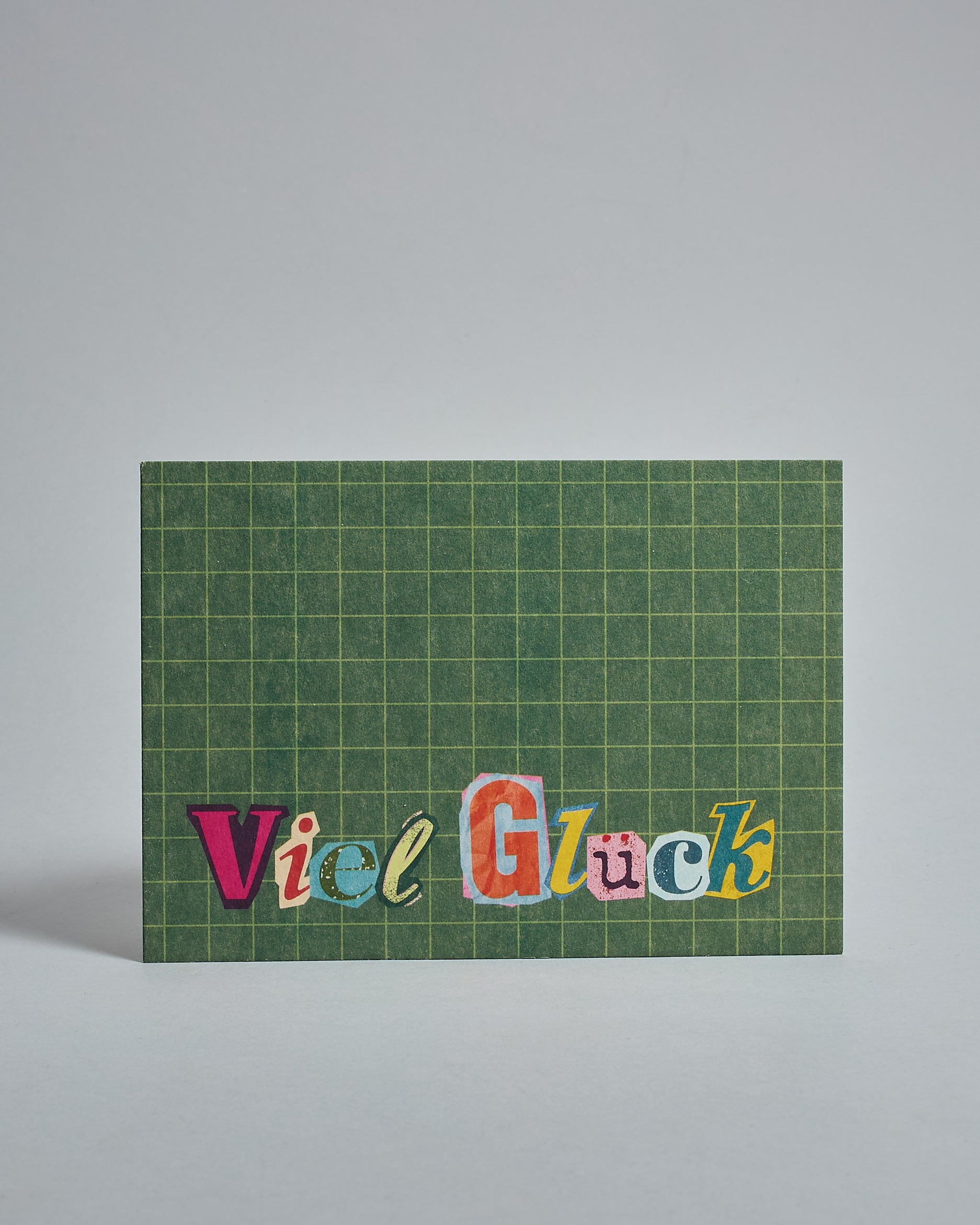 Die Vorderseite der Postkarte im Querformat vor hellgrauem Hintergrund. Im Vordergrund der Postkarte steht "Viel Glück" in Buchstaben die aussehen als wären sie aus Zeitrschriften ausgeschnitten. Der Hintergrund ist grasgrün mit dezentem Karomuster.