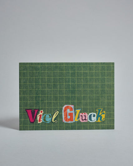 Die Vorderseite der Postkarte im Querformat vor hellgrauem Hintergrund. Im Vordergrund der Postkarte steht "Viel Glück" in Buchstaben die aussehen als wären sie aus Zeitrschriften ausgeschnitten. Der Hintergrund ist grasgrün mit dezentem Karomuster.