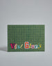 Die Vorderseite der Postkarte im Querformat vor hellgrauem Hintergrund. Im Vordergrund der Postkarte steht "Viel Glück" in Buchstaben die aussehen als wären sie aus Zeitrschriften ausgeschnitten. Der Hintergrund ist grasgrün mit dezentem Karomuster.