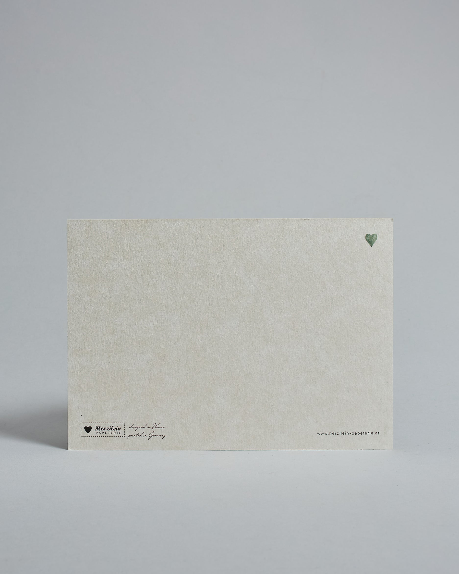 Die Rückseite der Postkarte im Querformat vor hellgrauem Hintergrund. Rechts oben ist, in kleiner Ausführung, ein grünes Herz zu sehen. 
Unten sind klein das Logo Herzilein-Papeterie, der Text: "designed in VIenna, printed in Germany" und "www.herzilein-papeterie.at" zu sehen. 
Der Großteil ist unbedruckt die Struktur der hochwertigen Holzschliffpappe ist gut zu sehen. Hier ist viel Platz zum Schreiben.