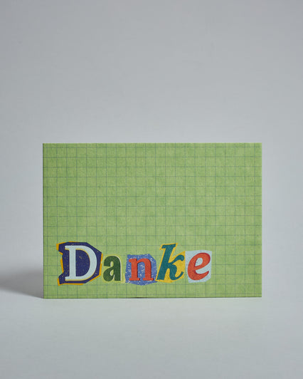 Die Vorderseite der Postkarte im Querformat vor hellgrauem Hintergrund. Im Vordergrund der Postkarte steht "Danke" in Buchstaben die aussehen als wären sie aus Zeitrschriften ausgeschnitten. Der Hintergrund ist hellgrün mit dezentem Karomuster.