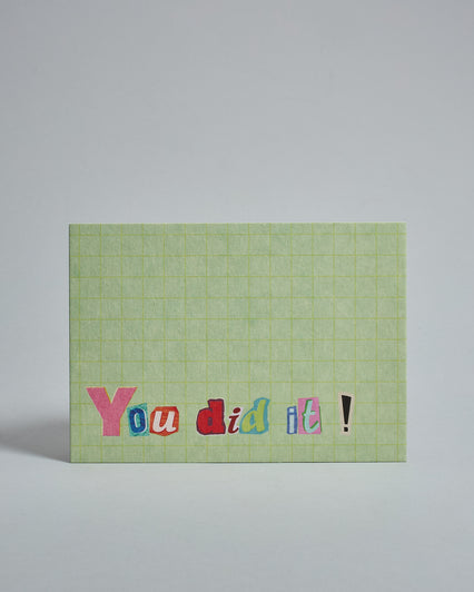 Die Vorderseite der Postkarte im Querformat vor hellgrauem Hintergrund. Im Vordergrund der Postkarte steht "You did it!" in Buchstaben die aussehen als wären sie aus Zeitrschriften ausgeschnitten. Der Hintergrund ist hellgrün mit dezentem Karomuster.