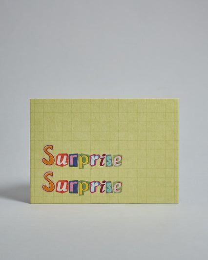 Die Vorderseite der Postkarte im Querformat vor hellgrauem Hintergrund. Im Vordergrund der Postkarte steht "Surprise Surprise" in zwei Zeilen in Buchstaben die aussehen als wären sie aus Zeitrschriften ausgeschnitten. Der Hintergrund ist grün-gelb mit dezentem Karomuster.