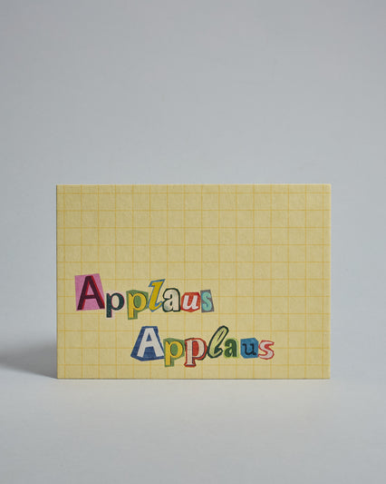 Die Vorderseite der Postkarte im Querformat vor hellgrauem Hintergrund. Im Vordergrund der Postkarte steht "Applaus Applaus" in zwei Zeilen in Buchstaben die aussehen als wären sie aus Zeitrschriften ausgeschnitten. Der Hintergrund ist gelb mit dezentem Karomuster.