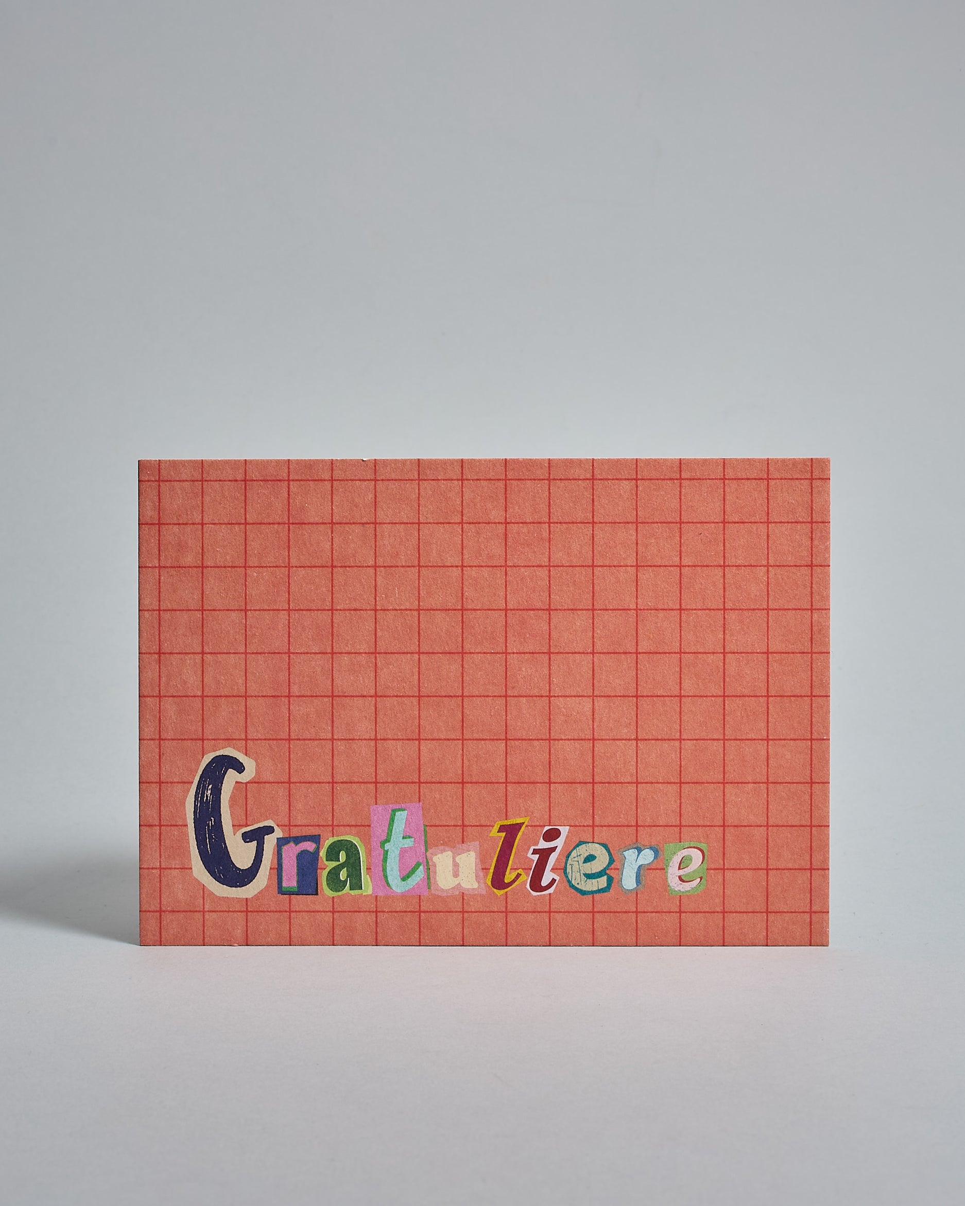 Die Vorderseite der Postkarte im Querformat vor hellgrauem Hintergrund. Im Vordergrund der Postkarte steht "Gratuliere" in Buchstaben die aussehen als wären sie aus Zeitrschriften ausgeschnitten. Der Hintergrund ist prange mit dezentem Karomuster.