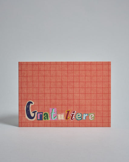 Die Vorderseite der Postkarte im Querformat vor hellgrauem Hintergrund. Im Vordergrund der Postkarte steht "Gratuliere" in Buchstaben die aussehen als wären sie aus Zeitrschriften ausgeschnitten. Der Hintergrund ist prange mit dezentem Karomuster.