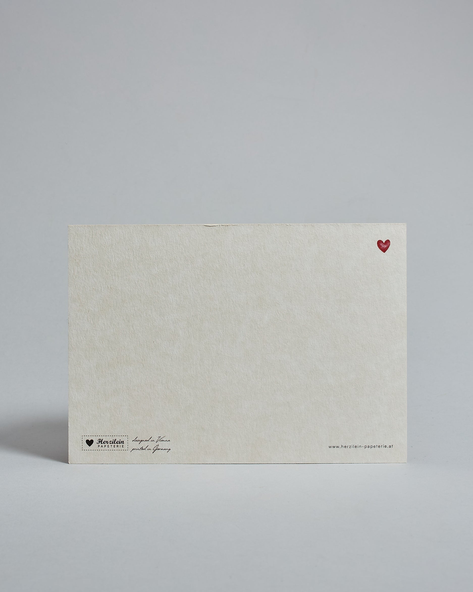 Die Rückseite der Postkarte im Querformat vor hellgrauem Hintergrund. Rechts oben ist, in kleiner Ausführung, ein rotes Herz zu sehen. 
Unten sind klein das Logo Herzilein-Papeterie, der Text: "designed in VIenna, printed in Germany" und "www.herzilein-papeterie.at" zu sehen. 
Der Großteil ist unbedruckt die Struktur der hochwertigen Holzschliffpappe ist gut zu sehen. Hier ist viel Platz zum Schreiben.