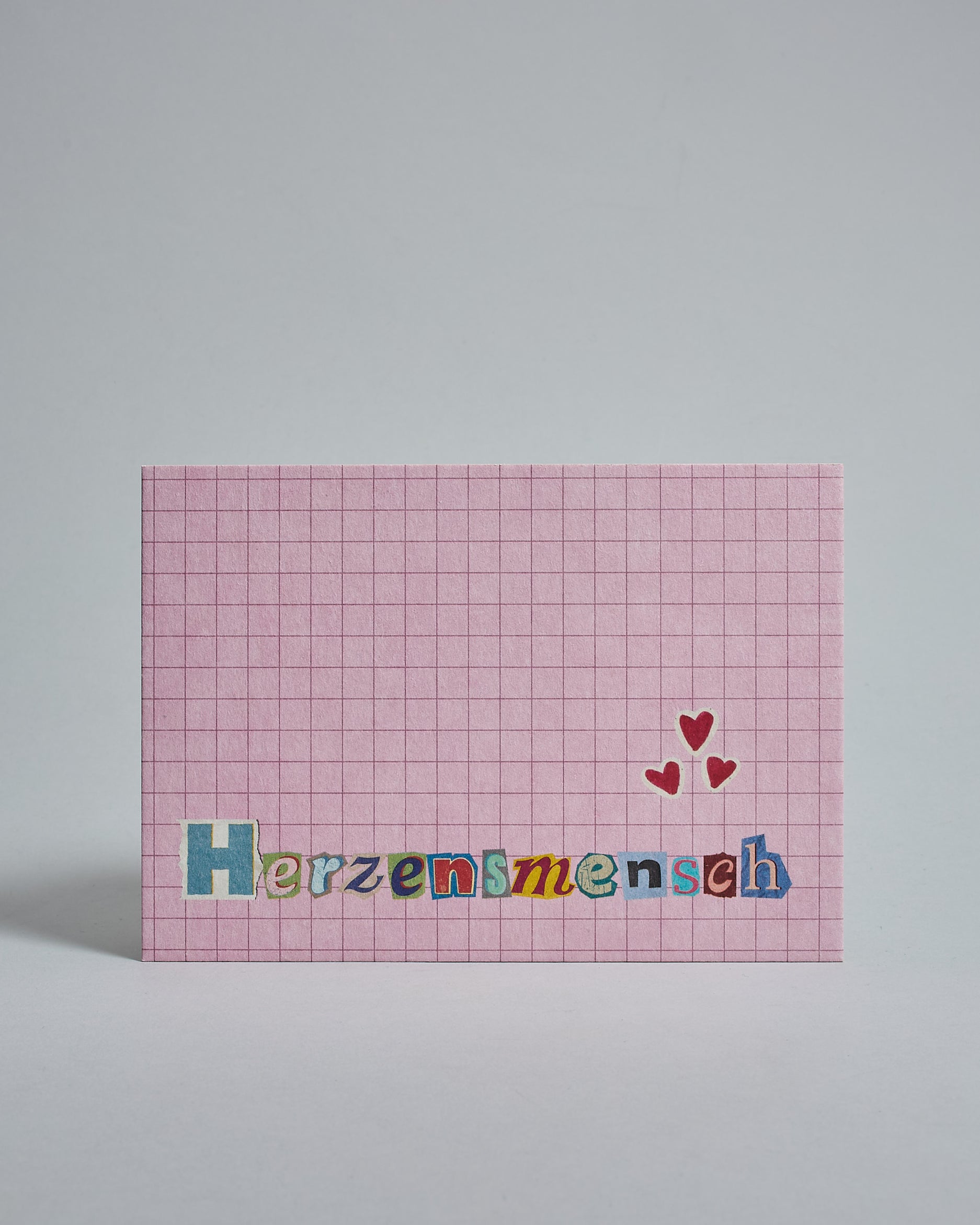 Die Vorderseite der Postkarte im Querformat vor hellgrauem Hintergrund. Im Vordergrund der Postkarte steht "Herzensmensch" in Buchstaben die aussehen als wären sie aus Zeitrschriften ausgeschnitten. Der Hintergrund ist rosa mit dezentem Karomuster.