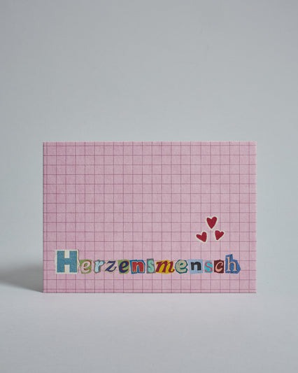 Die Vorderseite der Postkarte im Querformat vor hellgrauem Hintergrund. Im Vordergrund der Postkarte steht "Herzensmensch" in Buchstaben die aussehen als wären sie aus Zeitrschriften ausgeschnitten. Der Hintergrund ist rosa mit dezentem Karomuster.