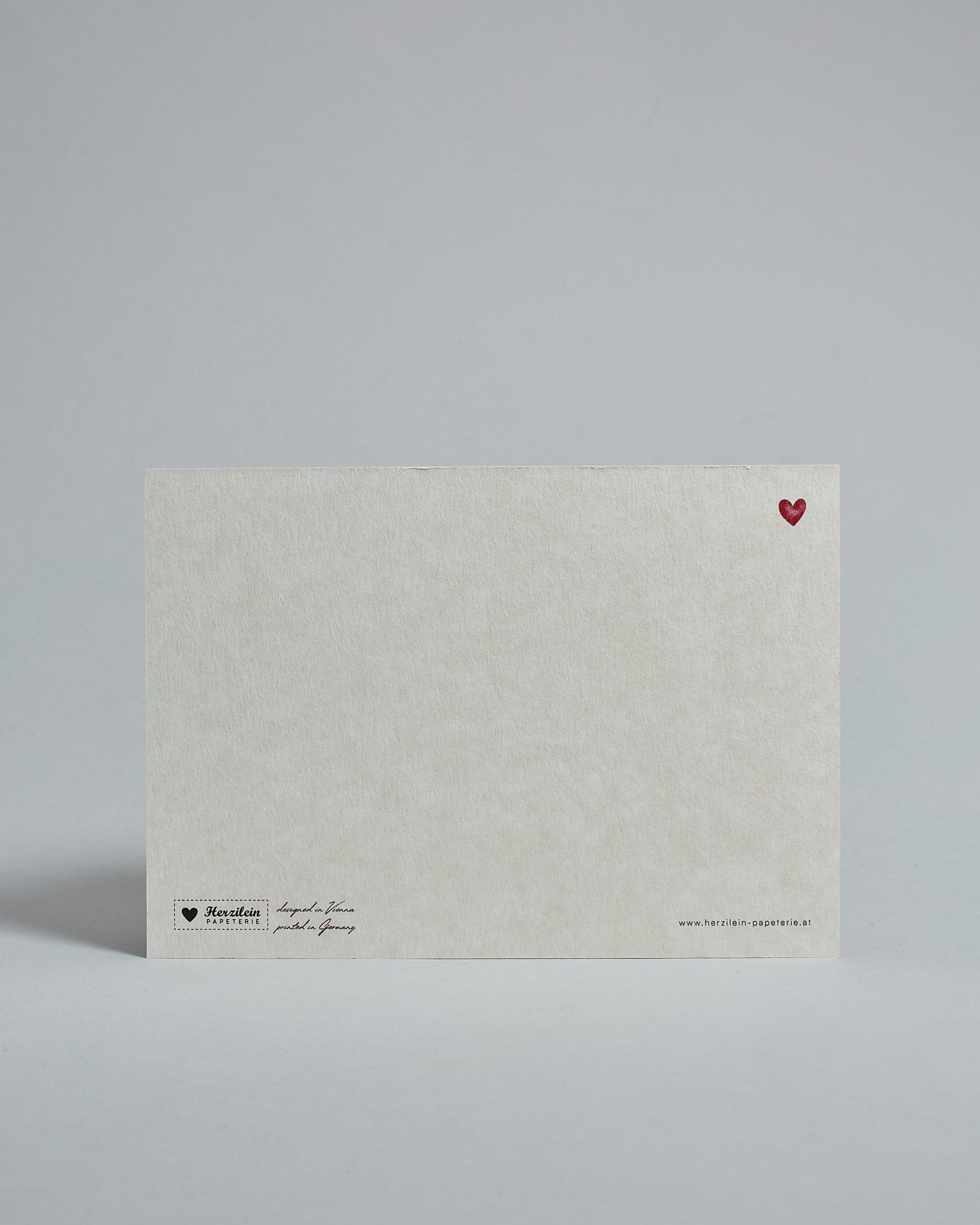 Die Rückseite der Postkarte im Querformat vor hellgrauem Hintergrund. Rechts oben ist, in kleiner Ausführung, ein rotes Herz zu sehen. 
Unten sind klein das Logo Herzilein-Papeterie, der Text: "designed in VIenna, printed in Germany" und "www.herzilein-papeterie.at" zu sehen. 
Der Großteil ist unbedruckt die Struktur der hochwertigen Holzschliffpappe ist gut zu sehen. Hier ist viel Platz zum Schreiben.