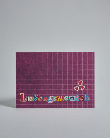 Die Vorderseite der Postkarte im Querformat vor hellgrauem Hintergrund. Im Vordergrund der Postkarte steht "Lieblingsmensch" in Buchstaben die aussehen als wären sie aus Zeitrschriften ausgeschnitten. Der Hintergrund ist weinrot mit dezentem Karomuster.