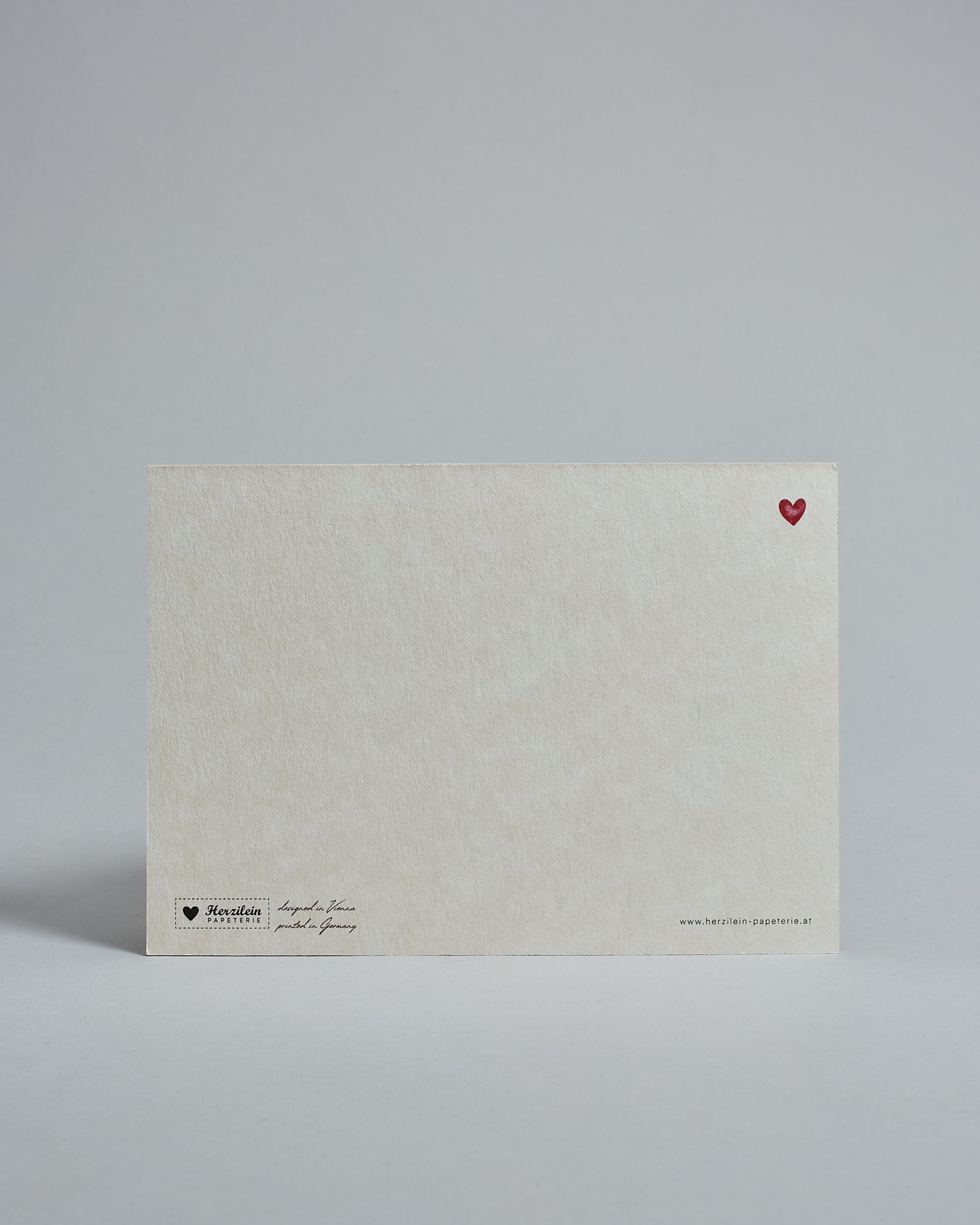 Die Rückseite der Postkarte im Querformat vor hellgrauem Hintergrund. Rechts oben ist, in kleiner Ausführung, ein rotes Herz zu sehen. 
Unten sind klein das Logo Herzilein-Papeterie, der Text: "designed in VIenna, printed in Germany" und "www.herzilein-papeterie.at" zu sehen. 
Der Großteil ist unbedruckt die Struktur der hochwertigen Holzschliffpappe ist gut zu sehen. Hier ist viel Platz zum Schreiben.
