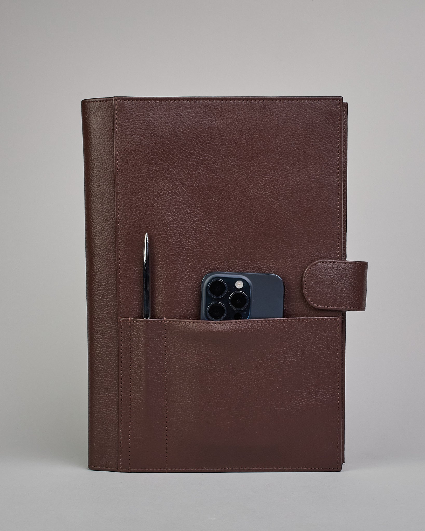 Dunkelbrauner Leder Notizbucheinband mit leichter Narbund. Der Einband hat außen ein flaches Fach in das ein Stift und das Handy gesteckt werden können. Das Notizbuch lässt sich seitlich mit einer Lasche und einem darunterliegenden Druckknopf verschließen. Innen bietet das Notizbuch rechts Platz um ein Buch einzustecken und links gibt es viele Fächer für weitere Notizzettel, Visitkarten und mehr. #4