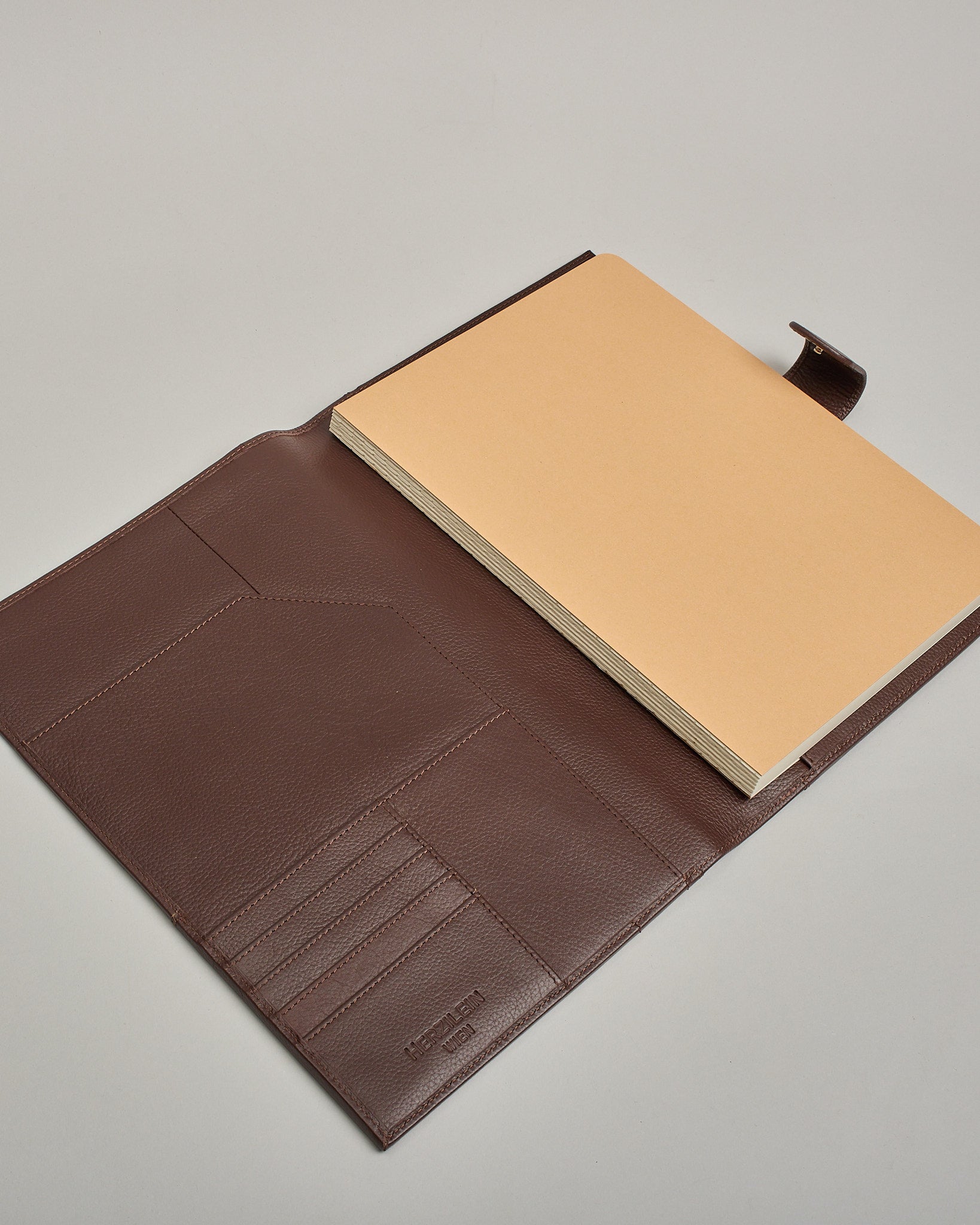 Dunkelbrauner Leder Notizbucheinband mit leichter Narbund. Der Einband hat außen ein flaches Fach in das ein Stift und das Handy gesteckt werden können. Das Notizbuch lässt sich seitlich mit einer Lasche und einem darunterliegenden Druckknopf verschließen. Innen bietet das Notizbuch rechts Platz um ein Buch einzustecken und links gibt es viele Fächer für weitere Notizzettel, Visitkarten und mehr. #7