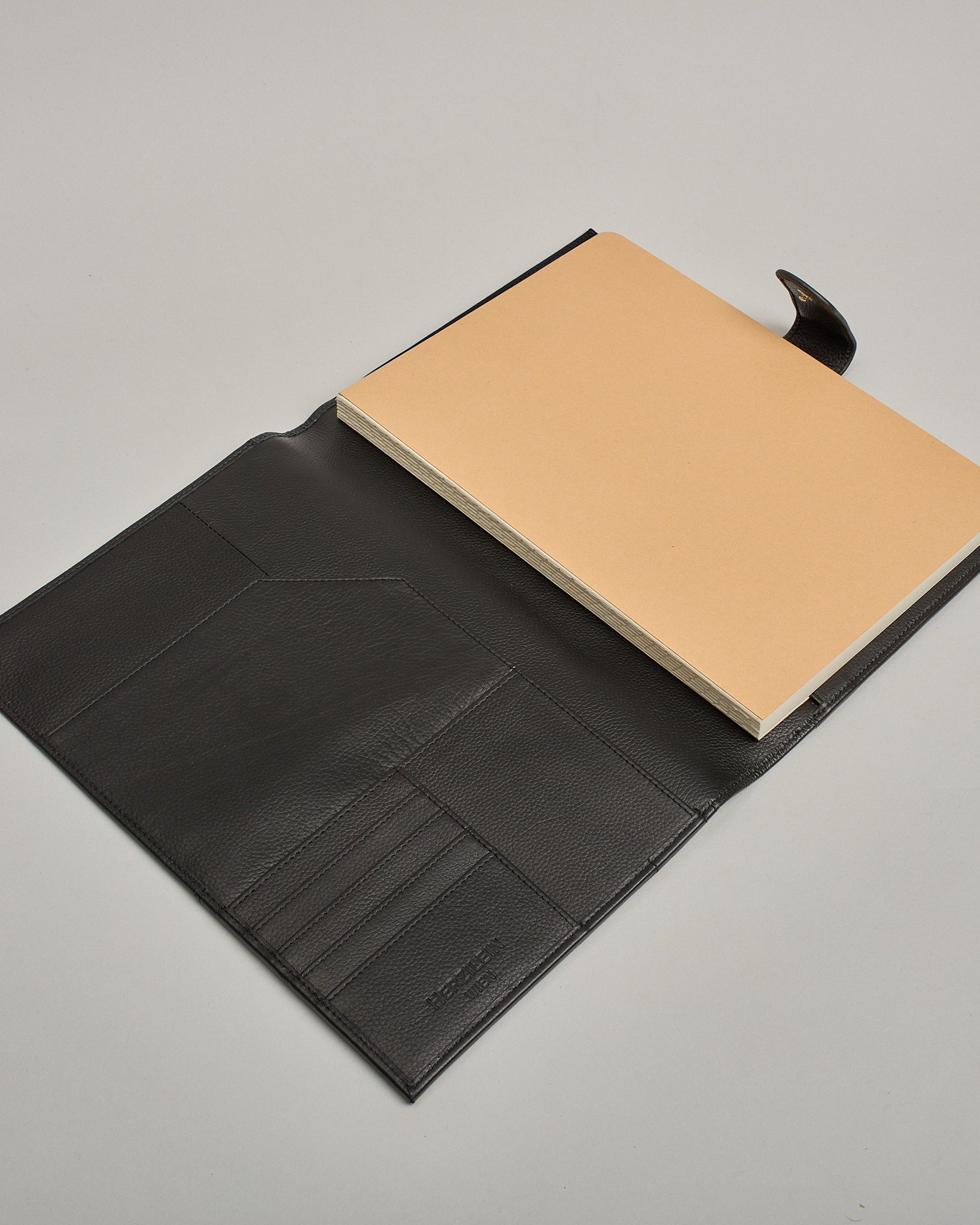 Schwarzer Leder Notizbucheinband mit leichter Narbund. Der Einband hat außen ein flaches Fach in das ein Stift und das Handy gesteckt werden können. Das Notizbuch lässt sich seitlich mit einer Lasche und einem darunterliegenden Druckknopf verschließen. Innen bietet das Notizbuch rechts Platz um ein Buch einzustecken und links gibt es viele Fächer für weitere Notizzettel, Visitkarten und mehr. #5