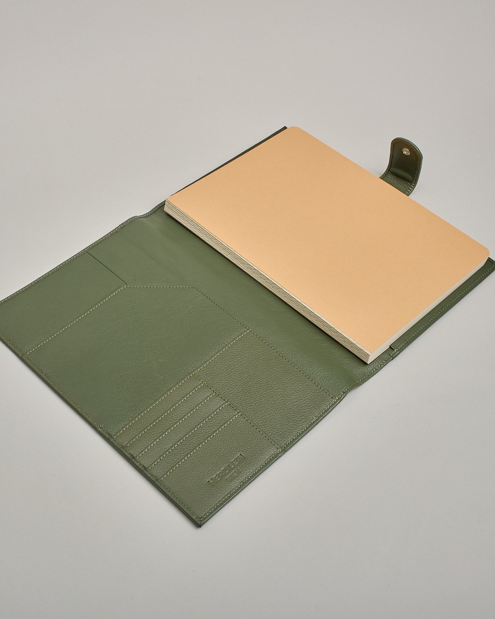 Dunkelgrüner Leder Notizbucheinband mit leichter Narbund. Der Einband hat außen ein flaches Fach in das ein Stift und das Handy gesteckt werden können. Das Notizbuch lässt sich seitlich mit einer Lasche und einem darunterliegenden Druckknopf verschließen. Innen bietet das Notizbuch rechts Platz um ein Buch einzustecken und links gibt es viele Fächer für weitere Notizzettel, Visitkarten und mehr. #5