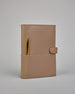 Beiger Leder Notizbucheinband mit leichter Narbund. Der Einband hat außen ein flaches Fach in das ein Stift und das Handy gesteckt werden können. Das Notizbuch lässt sich seitlich mit einer Lasche und einem darunterliegenden Druckknopf verschließen. Innen bietet das Notizbuch rechts Platz um ein Buch einzustecken und links gibt es viele Fächer für weitere Notizzettel, Visitkarten und mehr. #1
