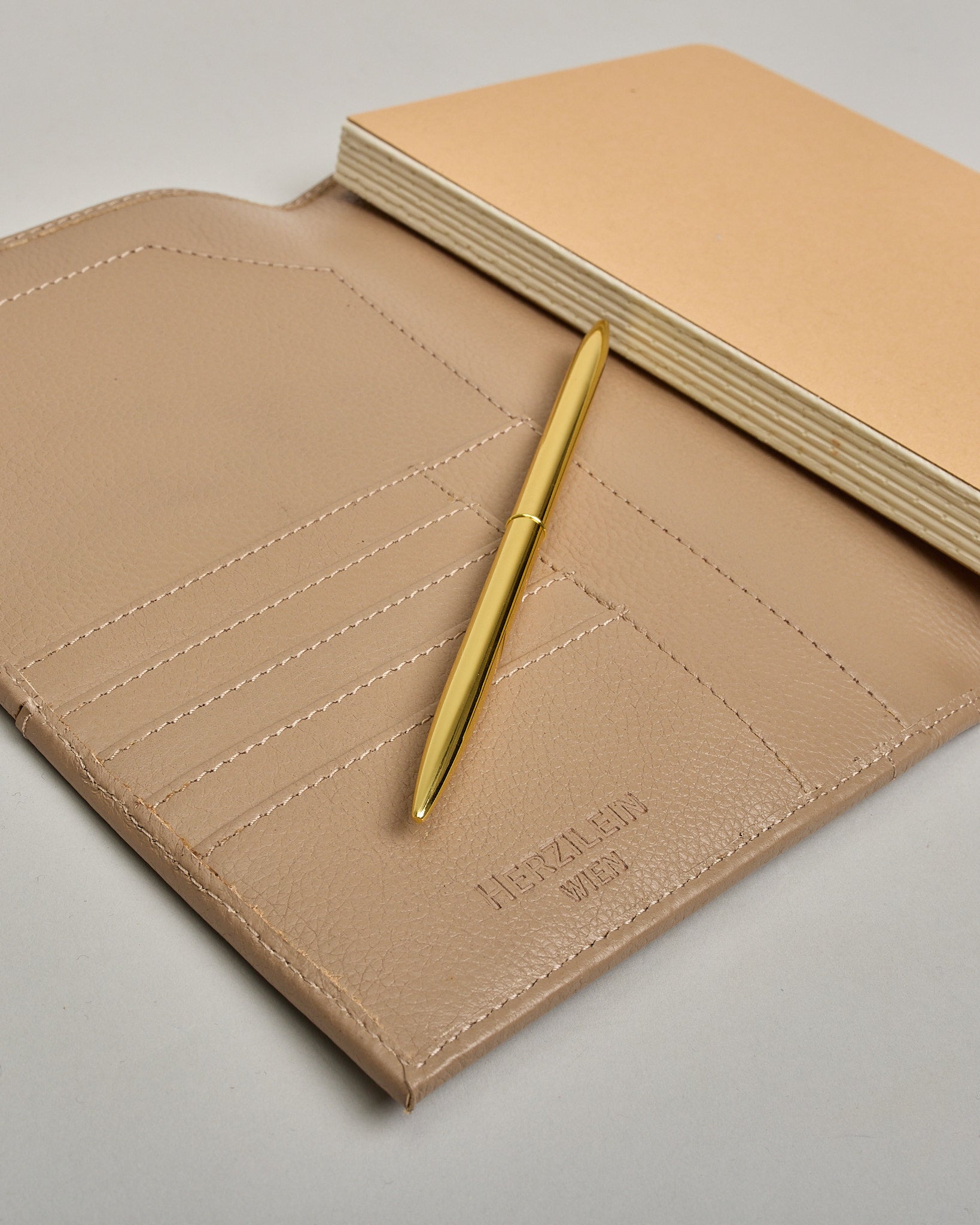 Beiger Leder Notizbucheinband mit leichter Narbund. Der Einband hat außen ein flaches Fach in das ein Stift und das Handy gesteckt werden können. Das Notizbuch lässt sich seitlich mit einer Lasche und einem darunterliegenden Druckknopf verschließen. Innen bietet das Notizbuch rechts Platz um ein Buch einzustecken und links gibt es viele Fächer für weitere Notizzettel, Visitkarten und mehr. #9