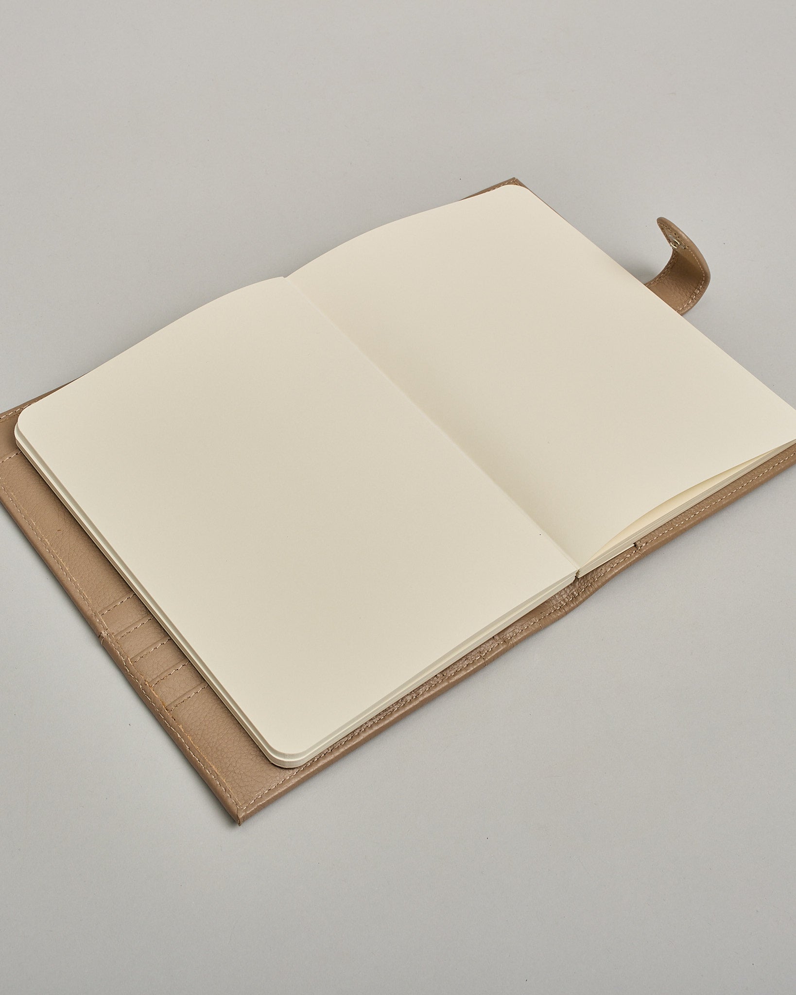 Beiger Leder Notizbucheinband mit leichter Narbund. Der Einband hat außen ein flaches Fach in das ein Stift und das Handy gesteckt werden können. Das Notizbuch lässt sich seitlich mit einer Lasche und einem darunterliegenden Druckknopf verschließen. Innen bietet das Notizbuch rechts Platz um ein Buch einzustecken und links gibt es viele Fächer für weitere Notizzettel, Visitkarten und mehr. #10