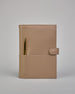 Beiger Leder Notizbucheinband mit leichter Narbund. Der Einband hat außen ein flaches Fach in das ein Stift und das Handy gesteckt werden können. Das Notizbuch lässt sich seitlich mit einer Lasche und einem darunterliegenden Druckknopf verschließen. Innen bietet das Notizbuch rechts Platz um ein Buch einzustecken und links gibt es viele Fächer für weitere Notizzettel, Visitkarten und mehr. #2