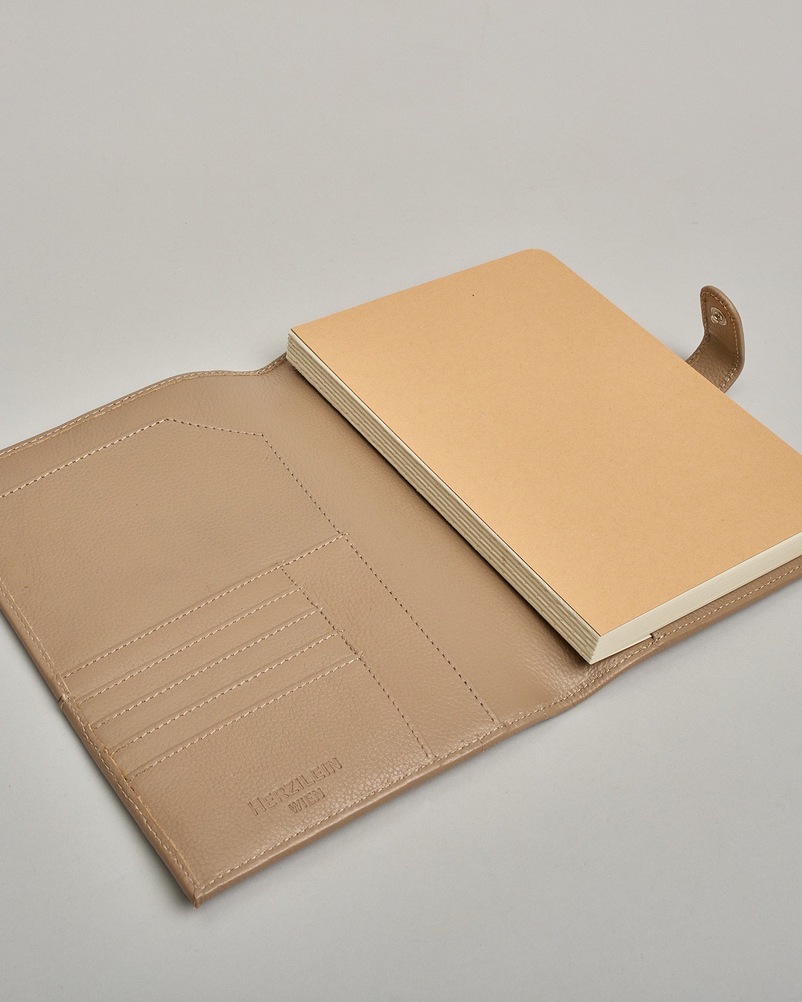 Beiger Leder Notizbucheinband mit leichter Narbund. Der Einband hat außen ein flaches Fach in das ein Stift und das Handy gesteckt werden können. Das Notizbuch lässt sich seitlich mit einer Lasche und einem darunterliegenden Druckknopf verschließen. Innen bietet das Notizbuch rechts Platz um ein Buch einzustecken und links gibt es viele Fächer für weitere Notizzettel, Visitkarten und mehr. #6