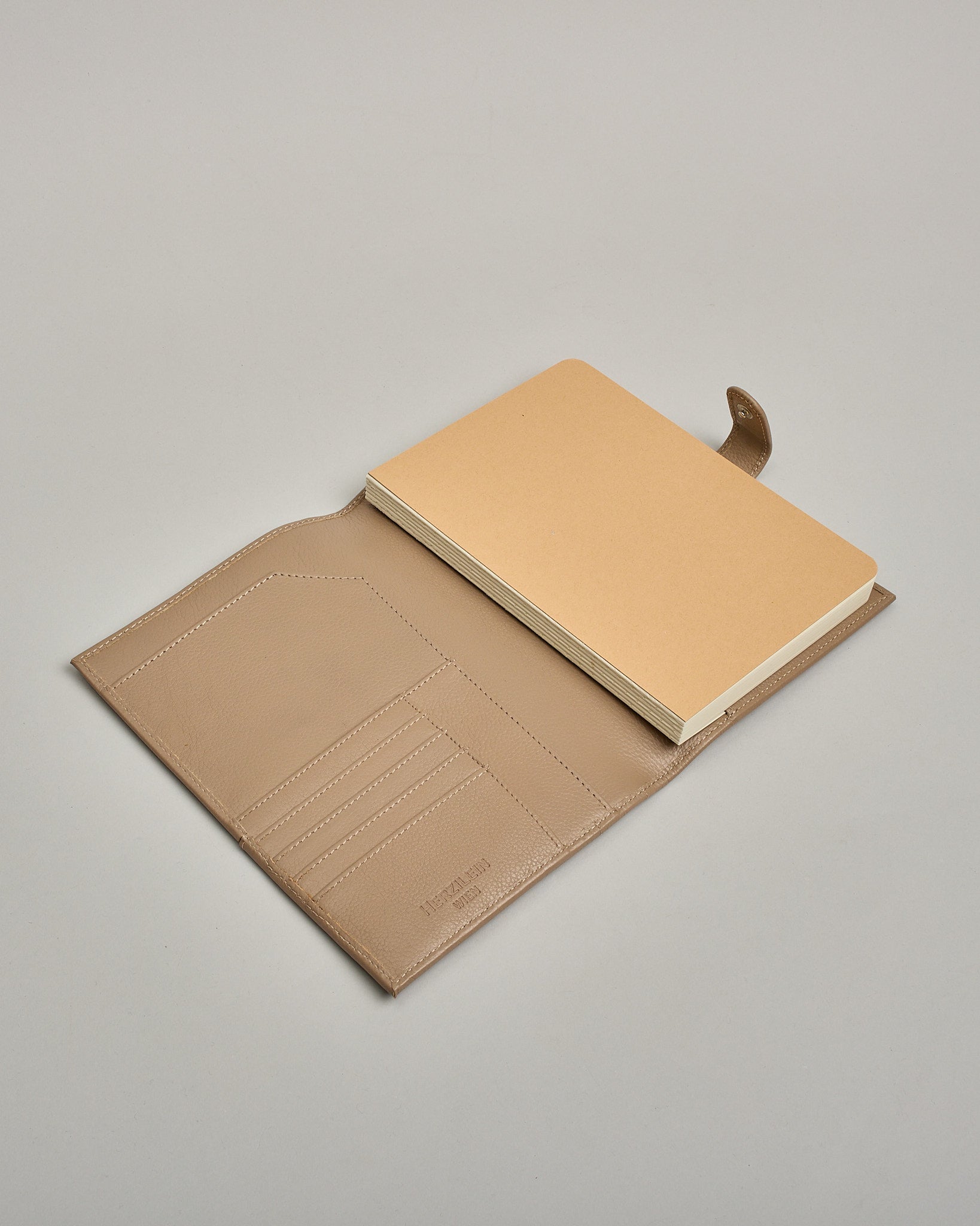 Beiger Leder Notizbucheinband mit leichter Narbund. Der Einband hat außen ein flaches Fach in das ein Stift und das Handy gesteckt werden können. Das Notizbuch lässt sich seitlich mit einer Lasche und einem darunterliegenden Druckknopf verschließen. Innen bietet das Notizbuch rechts Platz um ein Buch einzustecken und links gibt es viele Fächer für weitere Notizzettel, Visitkarten und mehr. #8