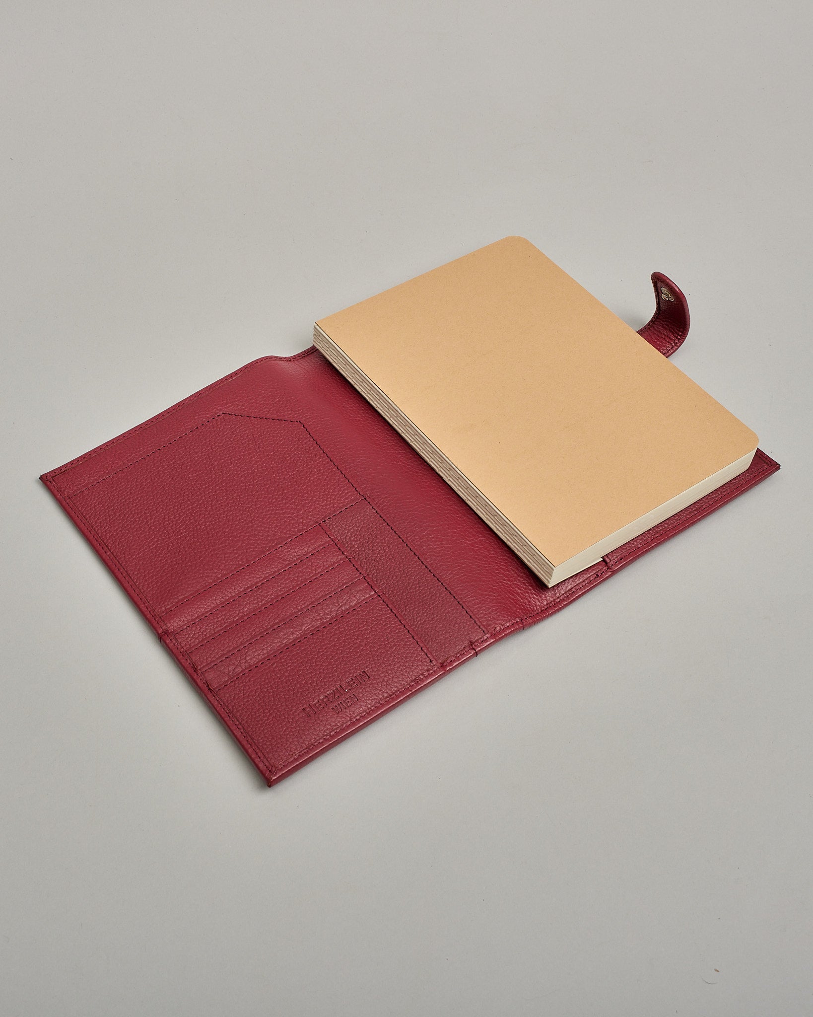 Bordeauxroter Leder Notizbucheinband mit leichter Narbund. Der Einband hat außen ein flaches Fach in das ein Stift und das Handy gesteckt werden können. Das Notizbuch lässt sich seitlich mit einer Lasche und einem darunterliegenden Druckknopf verschließen. Innen bietet das Notizbuch rechts Platz um ein Buch einzustecken und links gibt es viele Fächer für weitere Notizzettel, Visitkarten und mehr. #5